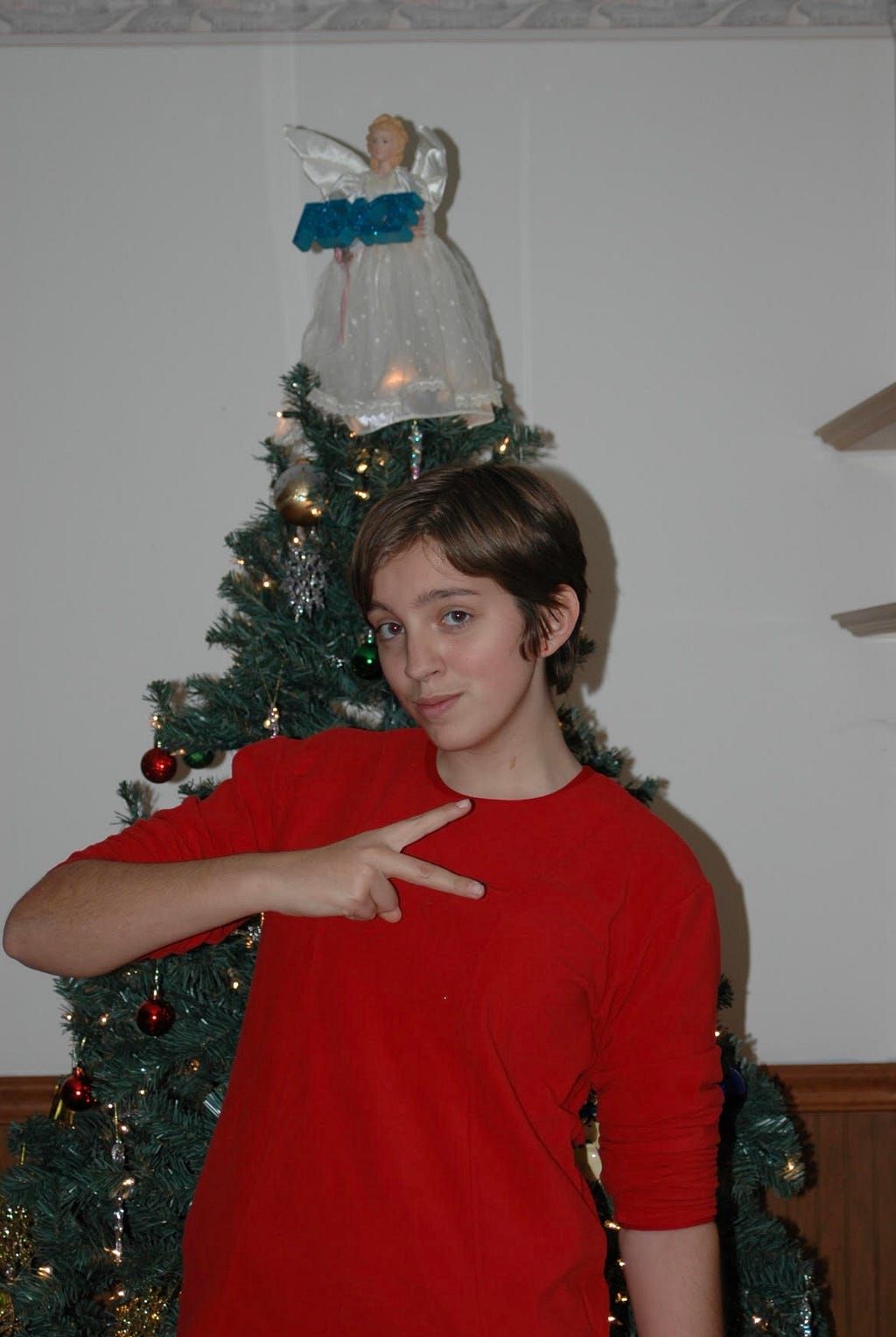A white female teenager of around fourteen looks into the camera while making a peace sign with her hands. She is wearing a red, long-sleeved shirt with the sleeves pushed up and there is a Christmas tree with an angel holding a sign which says “PEACE” in the background. The teenager has short brown hair.