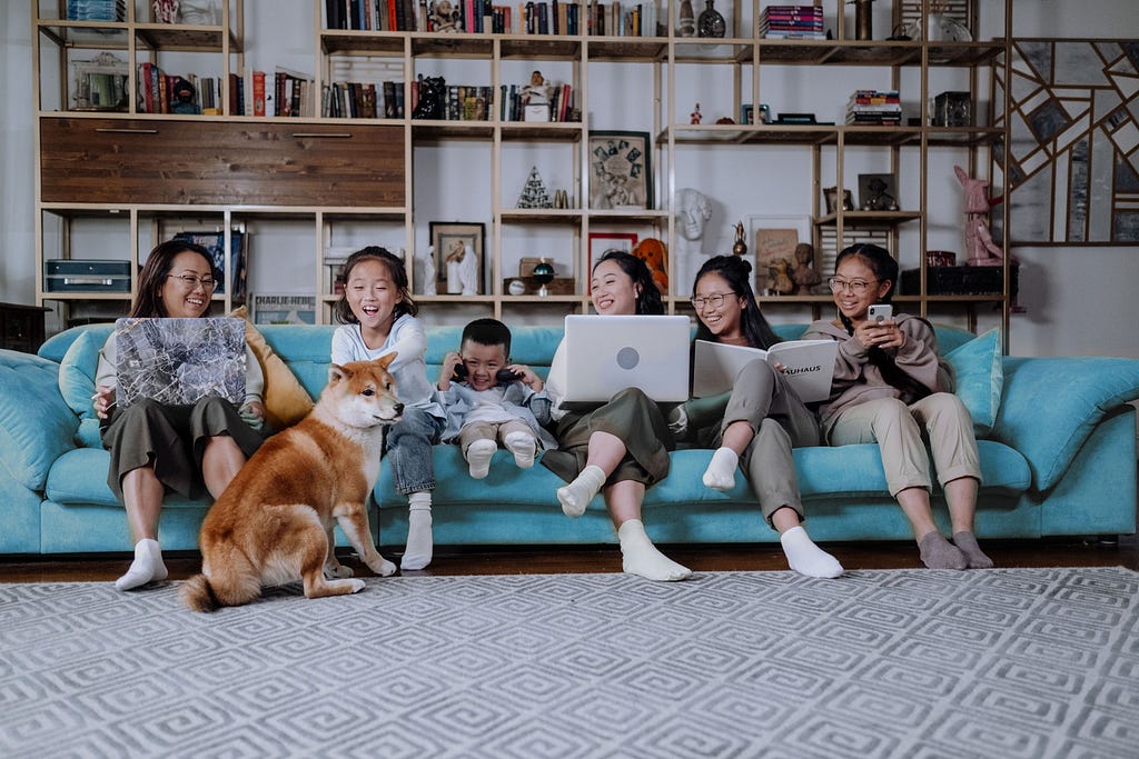 A family of six sit on a long couch on various internet enabled devices. They laugh at a dog on the floor.