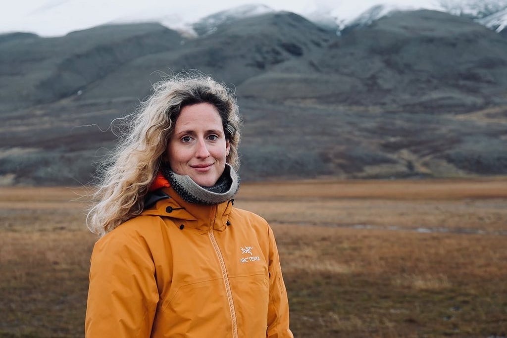 Maggie Coblentz faces the camera, wearing an orange winter jacket. Brown and grey snow-tipped mountains in the background.
