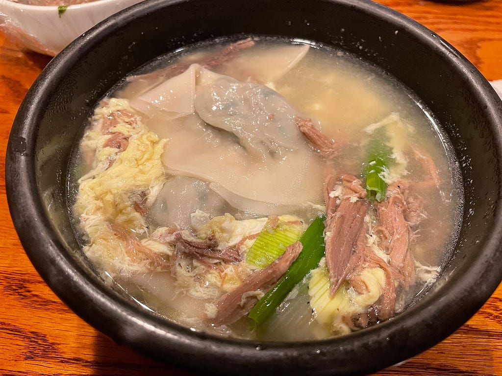 A black ceramic bowl that is filled with beef soup, egg drop, dumplings, sliced beef brisket, and green scallions