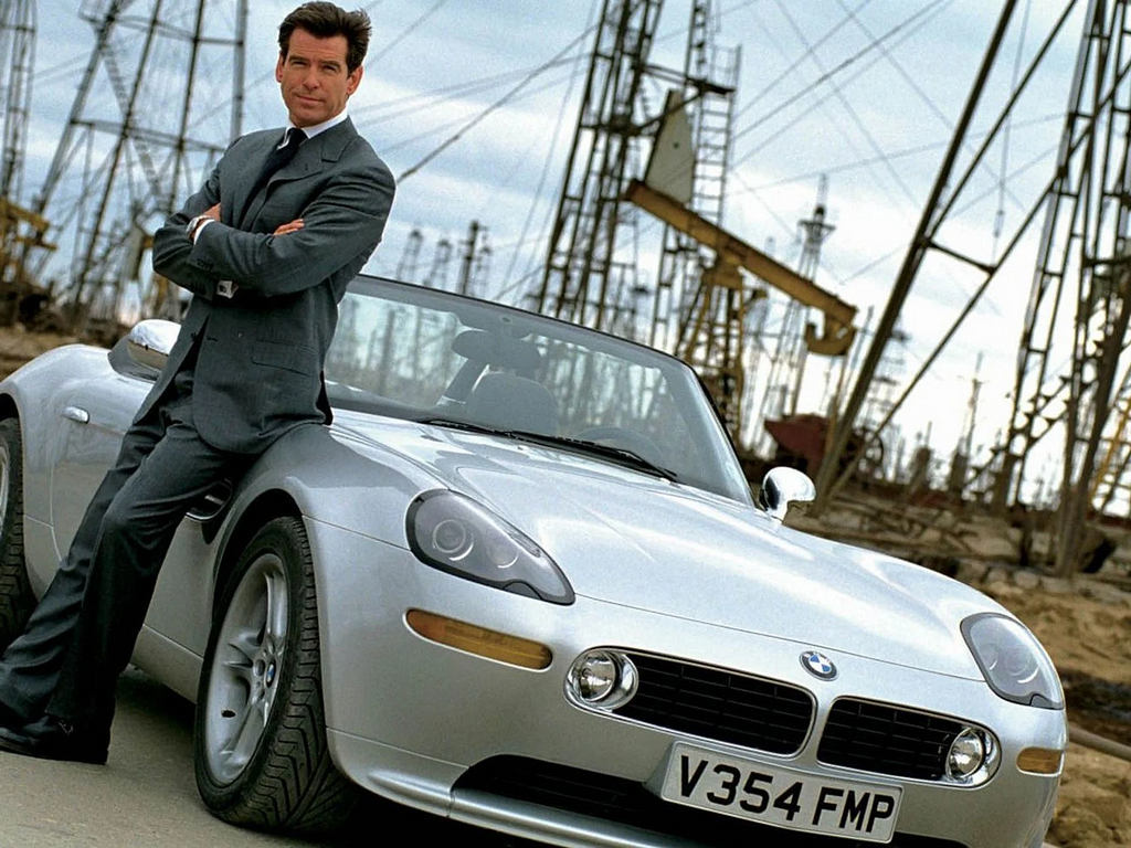 Pierce Brosnan, como James Bond, está parado al lado de un automóvil deportivo BMW Z8 plateado con la placa V354 FMP, posando con los brazos cruzados. El vehículo está estacionado en lo que parece ser una planta petrolífera, con varios pozos de petróleo visibles en el fondo.