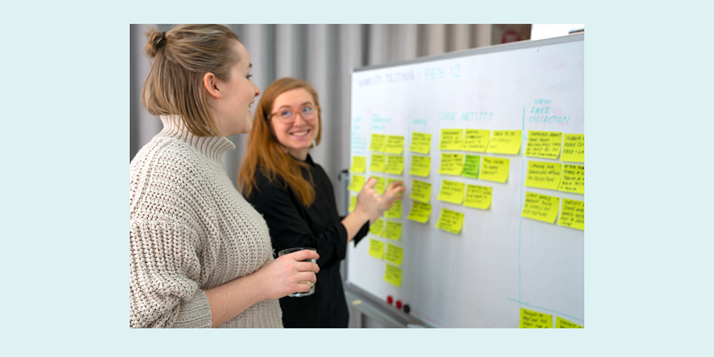 Two happy women discussing a whiteboard that is covered in sticky notes.