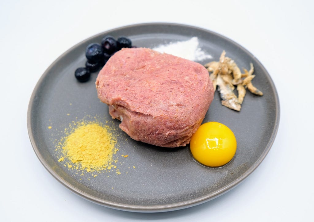 Raw food and add-ons displayed on a gray plate. Darwin’s raw cat food, with add-ins including egg yolk, nutritional yeast, blueberries, powdered goats milk, and freeze dried minnows (L to R). Full 8 oz. package — not a serving size suggestion.