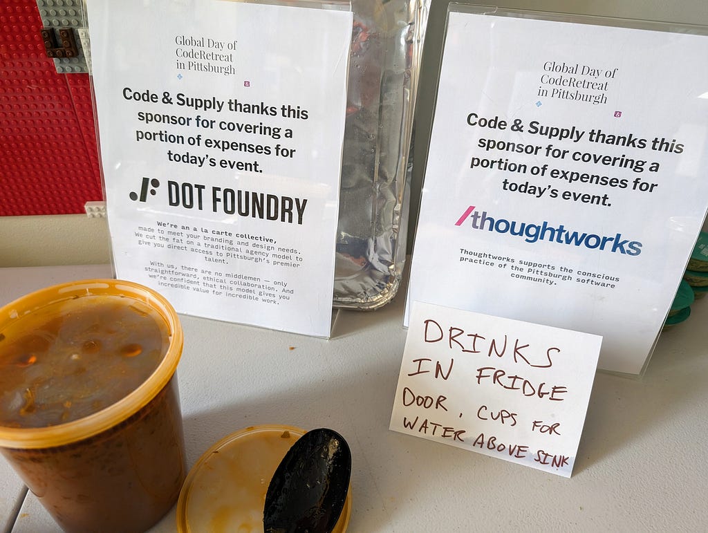 Two signs saying “Global Day of Coderetreat in Pittsburgh” and “Code & Supply thanks his sponsor for covering a portion of expenses for today’s event” with DOT FOUNDRY and THOUGHTWORKS on each of the two signs. Some illegible text is beneath the logos. The signs stand behind a container of food holding tikka masala. A sign in the foreground says DRINKS IN FRIDGE DOOR.