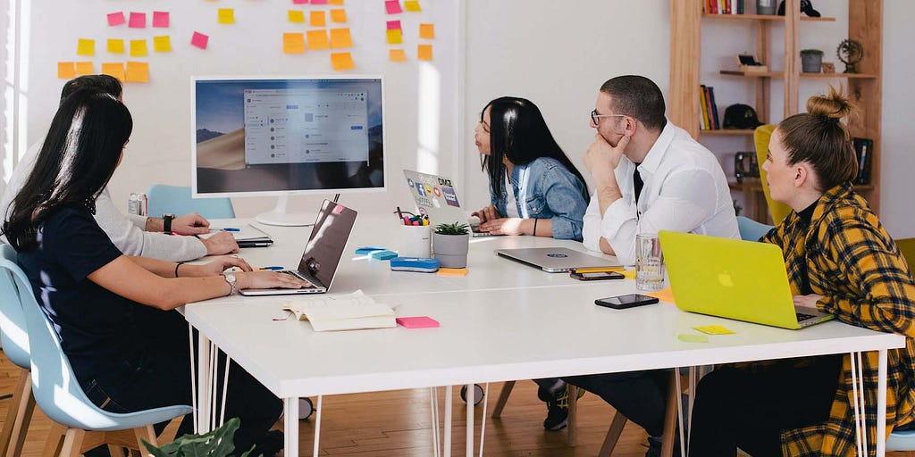 Photo of five employees sitting around a table with their laptops, looking at a computer monitor in the center. Post-it notes are on the wall behind them.