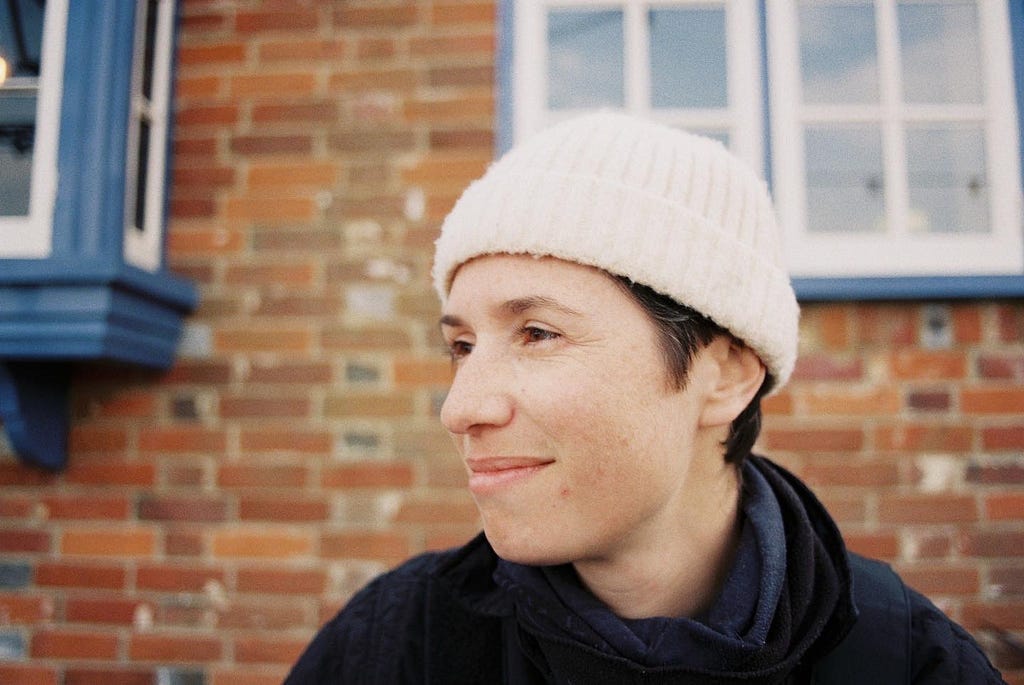 A picture of a person with white skin and a cream beanie hat in front of a red brick wall