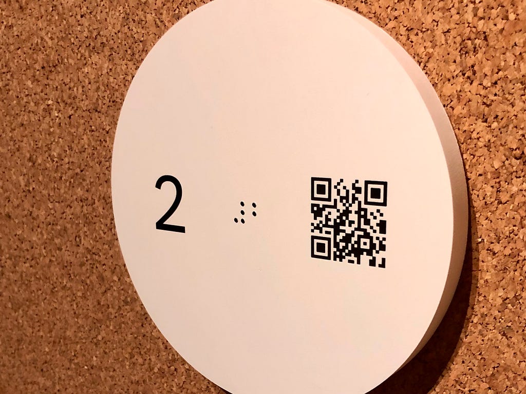 White disc on a cork wall, written on the disc is the number two written as a glyph, in brail and as a QR Code. The QR code when scanned with your phone will open a URL for the digital guide as stop 2
