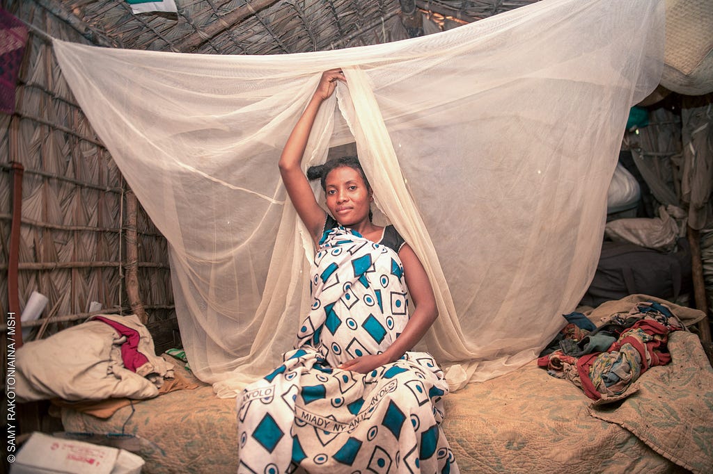 A pregnant woman sits on the edge of her bed under a mosquito net