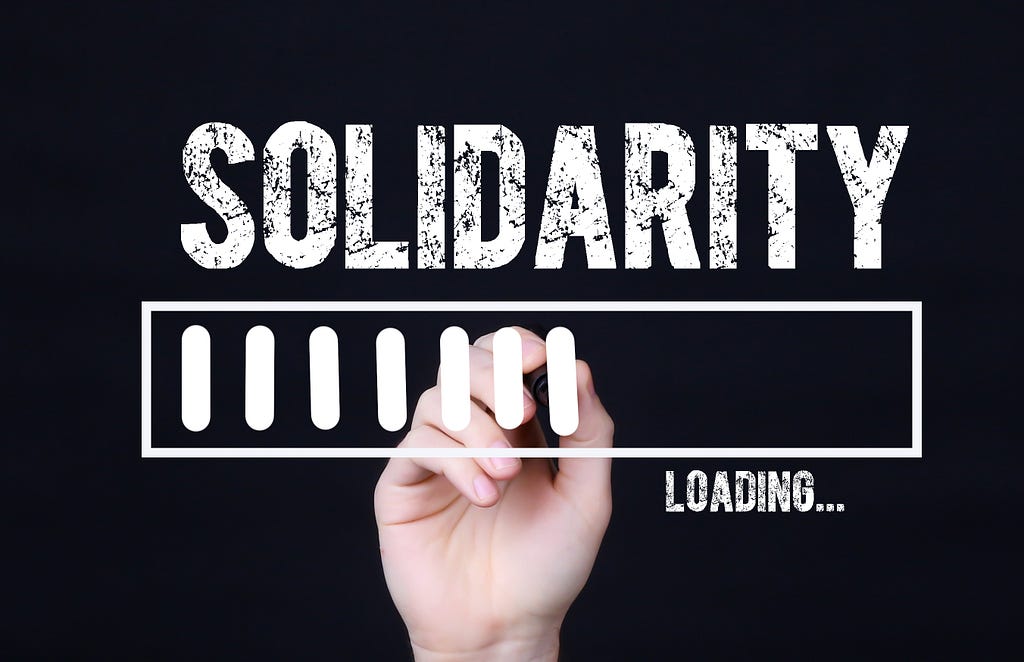 The word “Solidarity” in scratchy white block letters on a black background. Below the words is a thin, horizontal rectangle outlined in white with 7 thick, white tick marks meant to suggest an app loading. In small white letters beneath the rectangle is the word, “loading.” Behind the rectangle is a white, human hand that looks as if it’s adding the tick marks by hand.