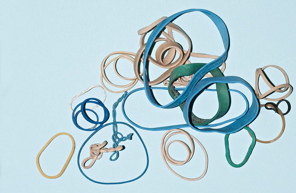 Lots of different circular rubber bands on a bright background thrown on top of each other. Some are twisted, others are nearly round.