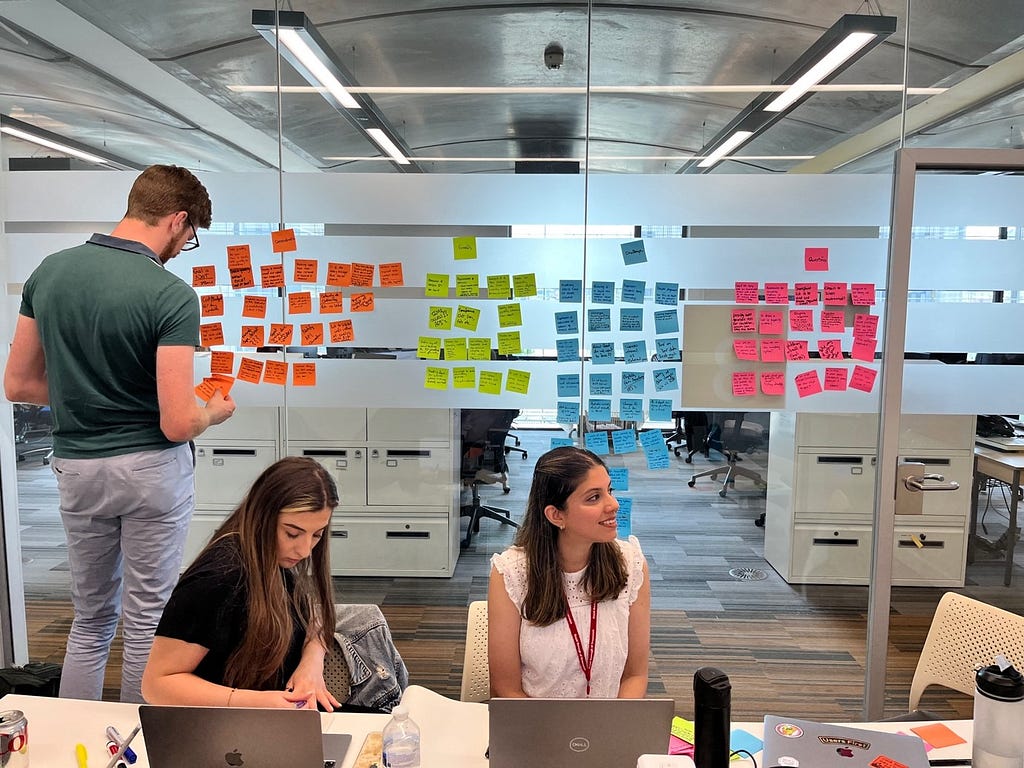 3 colleagues in a hackathon workshop. 1 is adding sticky notes to a glass wall, 2 sit at a table in front of laptops.