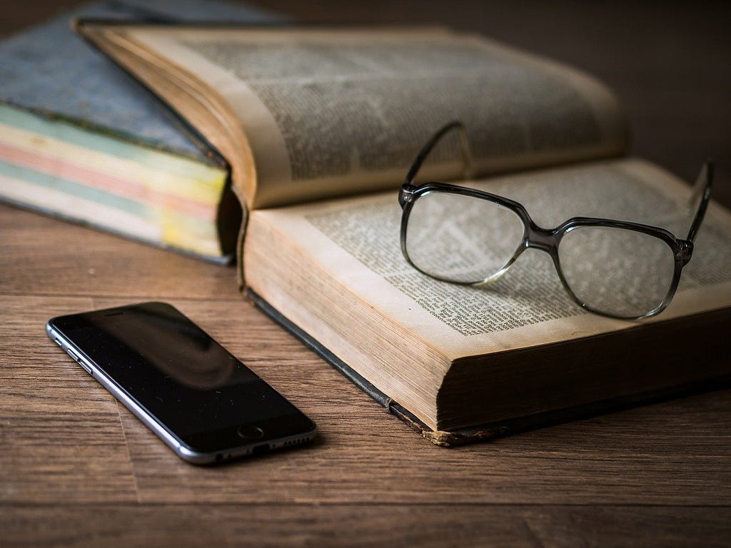 A mobile phone and two old novels on a wooden table. One novel is open with a pair of glasses on top the pages