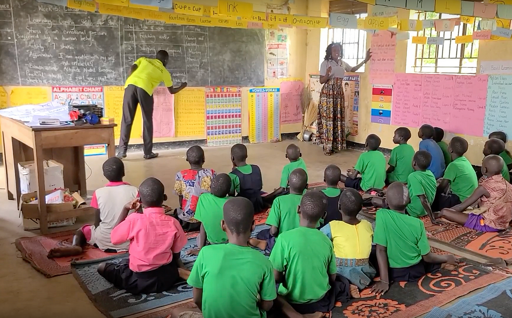 There’s a classroom with yellow walls, a blackboard and lots of colorful charts decorating the walls. A male teacher writes on the blackboard with his back to the camera, while a female teacher faces the learners. Learners are sat on the floor on yoga mats. They face the blackboard, their backs to the camera.