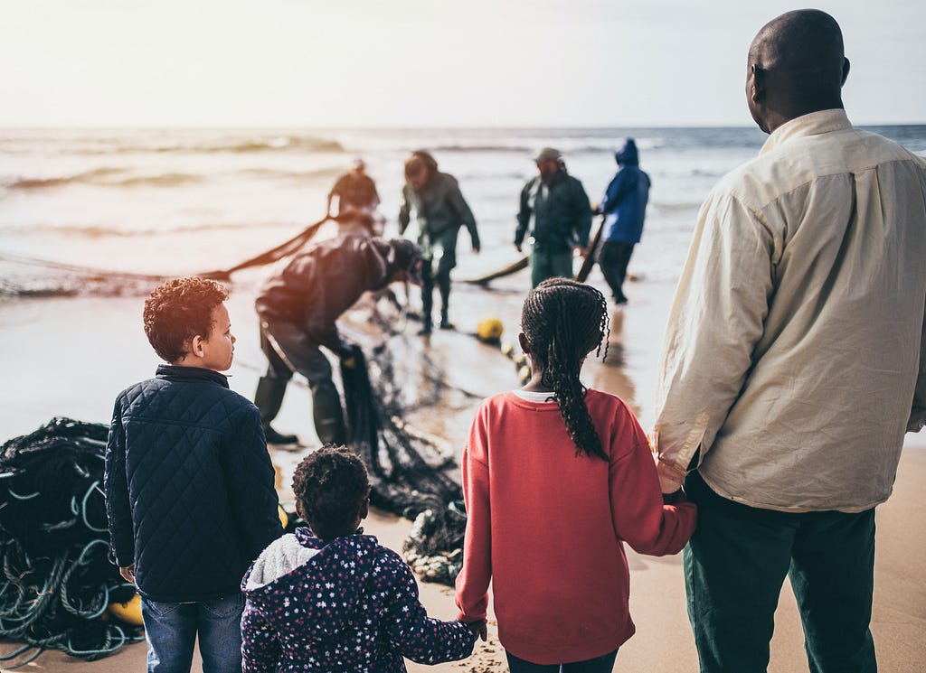 The front part of the image shows a group of people look afar, comprised of an adult and three children. They are holding hands, which suggests that they are a family. At the back, other individuals are seeing picking things from a fishnet at the seashore.