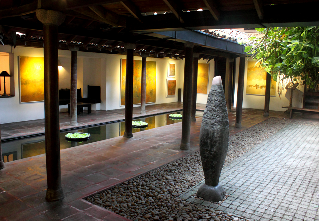The courtyard area of the Gallery Cafe with a rectangular pond in the middle with some water lettuce planted in 3 circular containers. There are corridors to walk around the pond, one side has walls and the other is open with polls supporting the roof. Walls are decorated with various paintings and the open side of the corridor connects to an open compound decorated with stones.