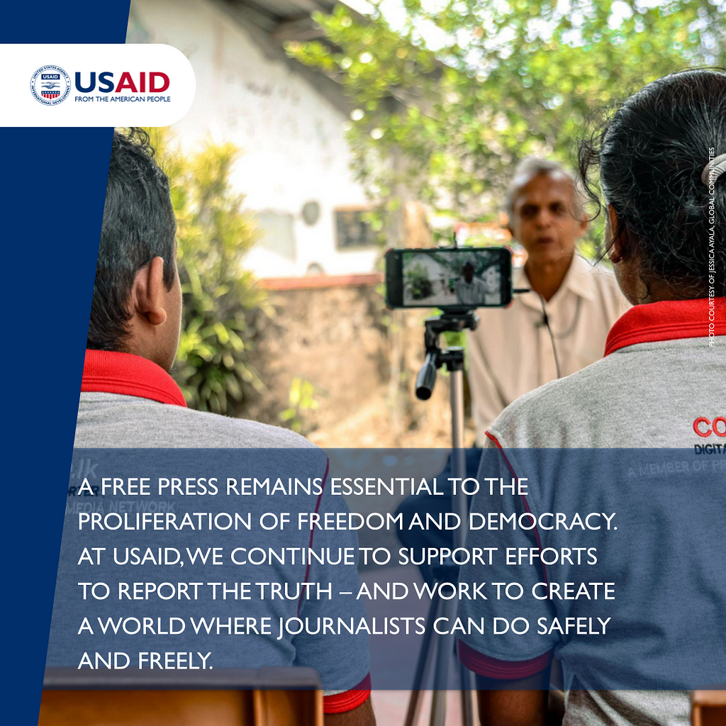 Two people wearing matching branded shirts that identify them as digital reporters sit side-by-side with their backs to the camera while interviewing a gray-haired man with a mobile phone perched on a tripod. A statement underneath the photo reads: A free press remains essential to the proliferation of freedom and democracy. At USAID, we continue to support efforts to report the truth — and work to create a world where journalists can do safely and freely.