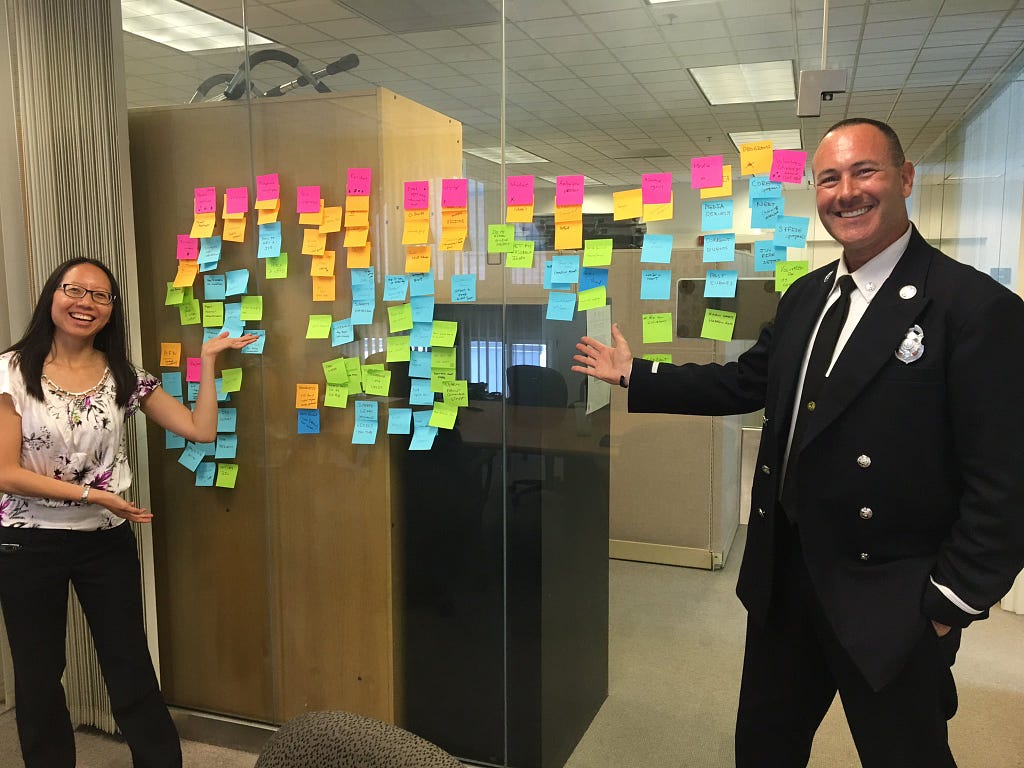 A content designer and Public Information Officer pose with a meeting room window covered in Post-Its after a workshop.