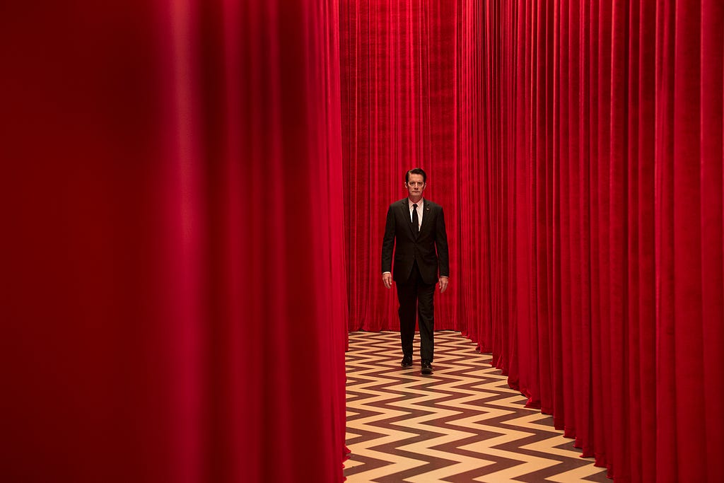 A still frame from Twin Peaks of a man in a suit walking on a zigzag floor surrounded by red curtains