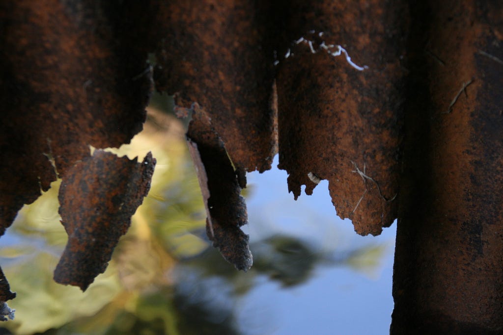 A close-up photo of rusted steel, pieces flaking off, in the background is water in which the reflection of sky and greenery is visible