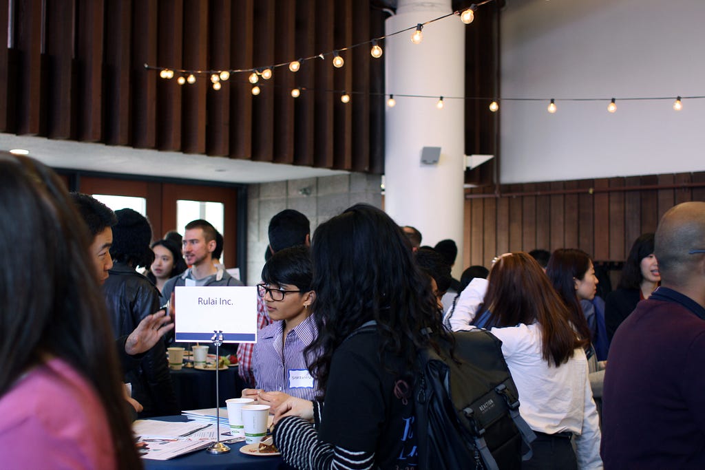 Photo of students speaking with employers and recruiters at a STEM employers event. People are gathered around tall tables indoors. There are lights strung from the ceiling.