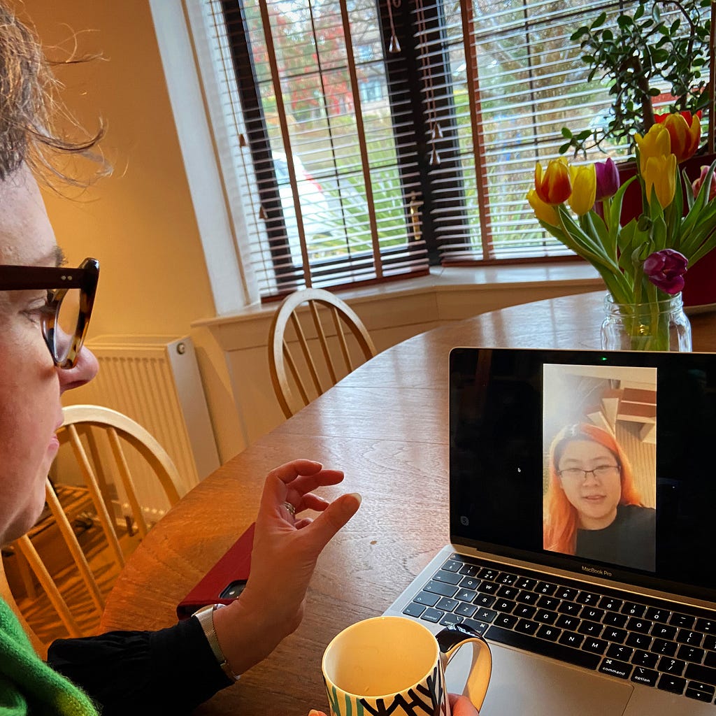 Hazel sitting at table with laptop talking to Jiaru who appears the laptop screen