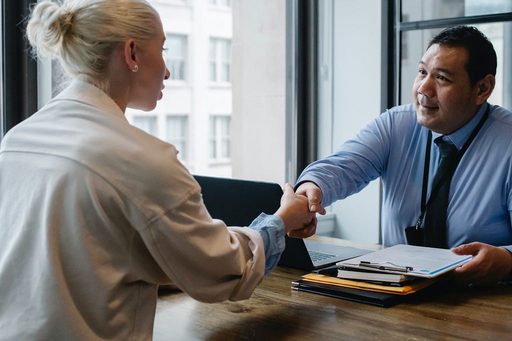 A picture of an Asian man (the interviewer) shaking a white woman’s hand at a job interview. Learn what questions to ask at the end of an interview.