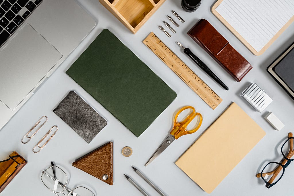 Tools such as a ruler, scissors, laptop, notebook, glasses, pen, and paper are laid out in a ‘flatlay’ shot, and it’s styled in a way which makes it organized and pleasing to look at.