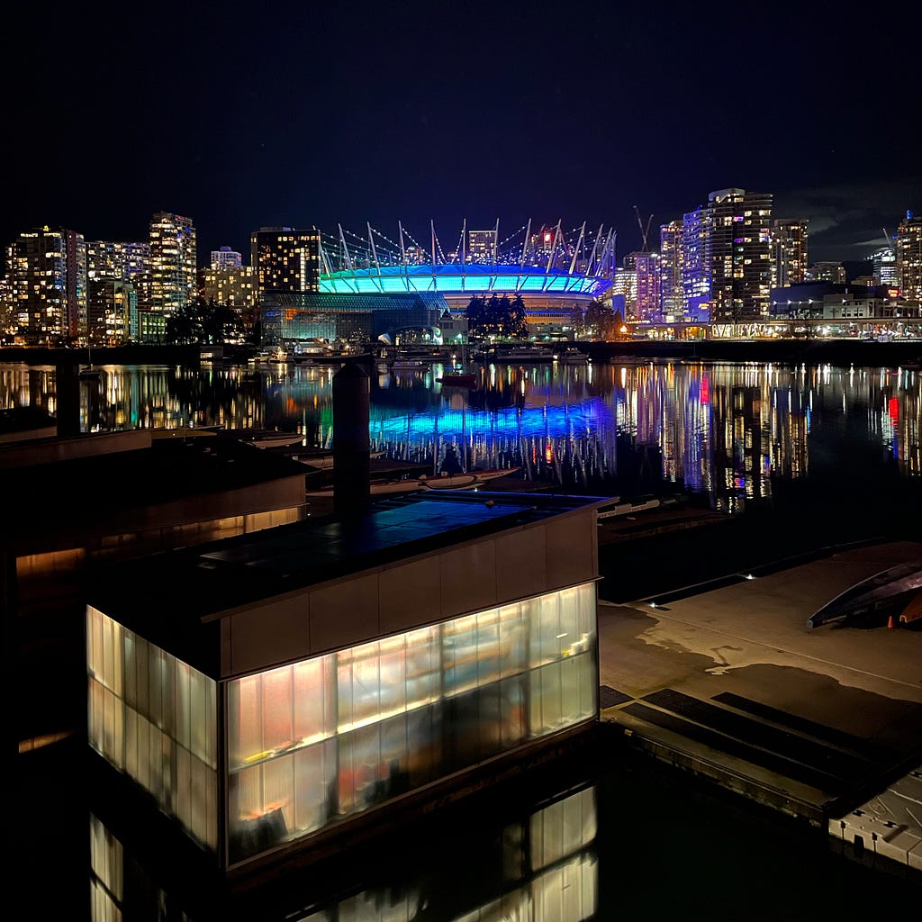 A cityscape at night, showing a lit stadium, hi-rises, and a body of water