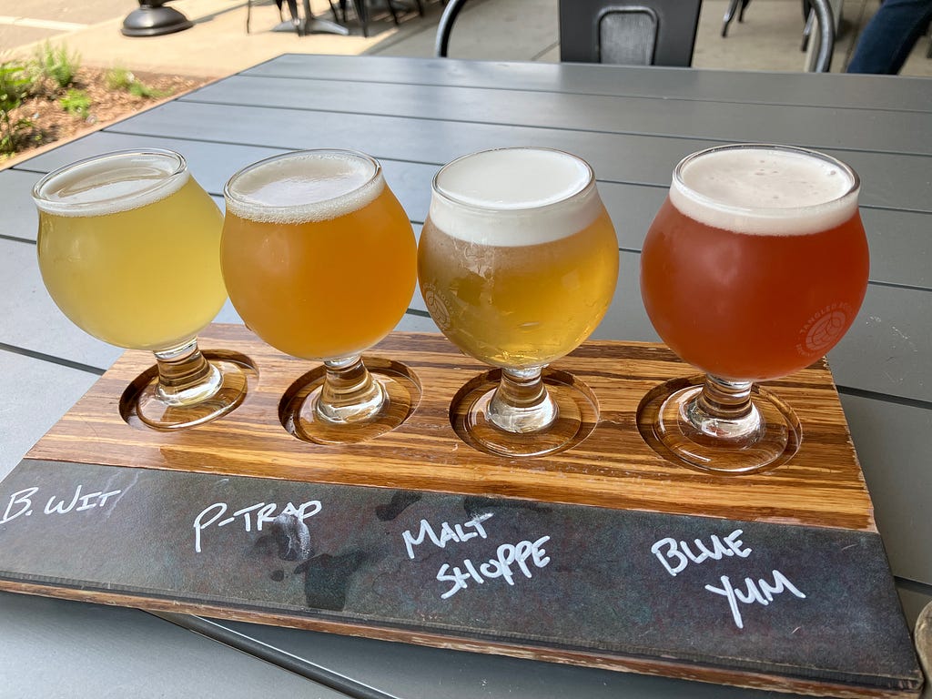 A flight of four beers on a wooden tray on an outdoor table, with labels containing the name of each beer.