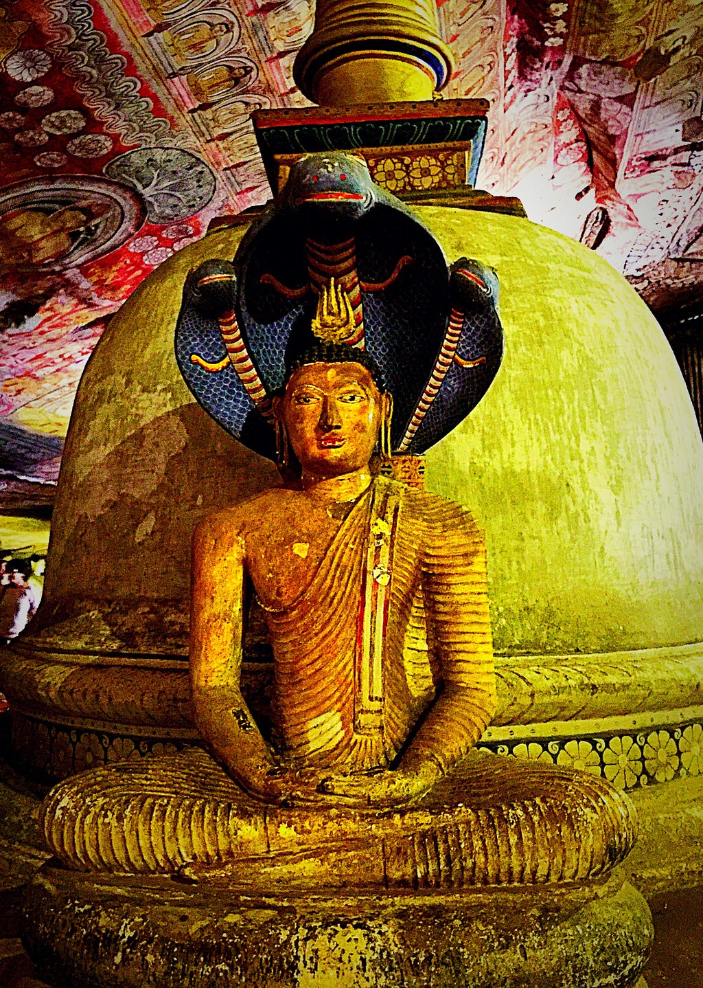 A photograph of a color painted, wooden statue of the Lord Buddha, seated, in front of a kovil (looks like a German WW I helmet, but it’s a temple. Behind the Buddha, is a giant cobra, flaring it’s hood above his head.