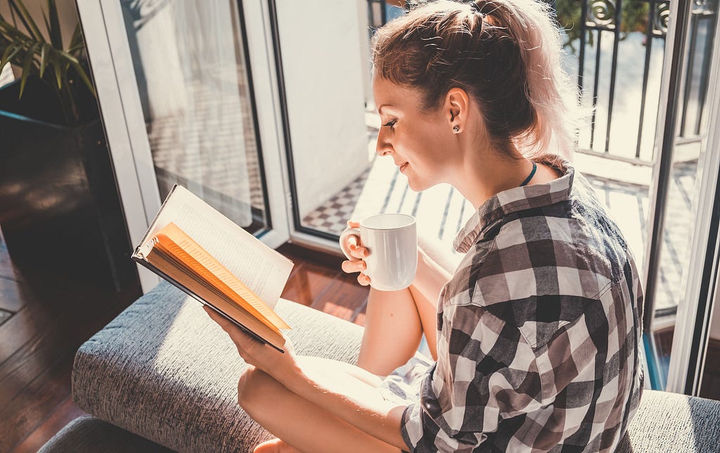 A woman sitting at opened window in a naturally lit room, drinking coffee and reading a book.