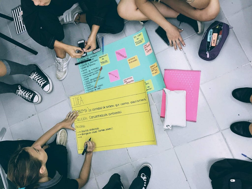 Young people stand around posters with ideas for deplastification. One poster is converted in post-it notes. They sit in a circle, some converses visible in the frame.