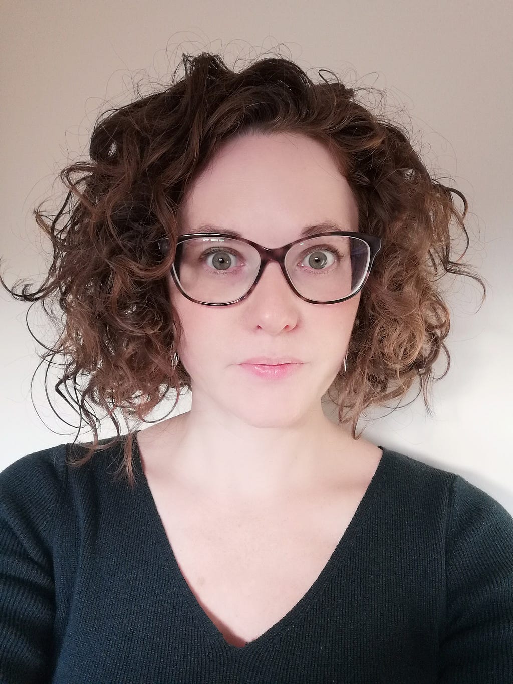 A serious-looking white woman wearing brown glasses with curly brown hair and green eyes. She is wearing a green v-neck top.