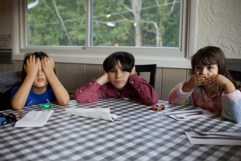 We've got three children sitting in the dinner table doing homework. From left to right, one is folding their eyes with their hands, the middle one is taping his ears with his hands and the last one is covering her mouth with her hands. There's a window on the back showing threes on the outside. It's afternoon.