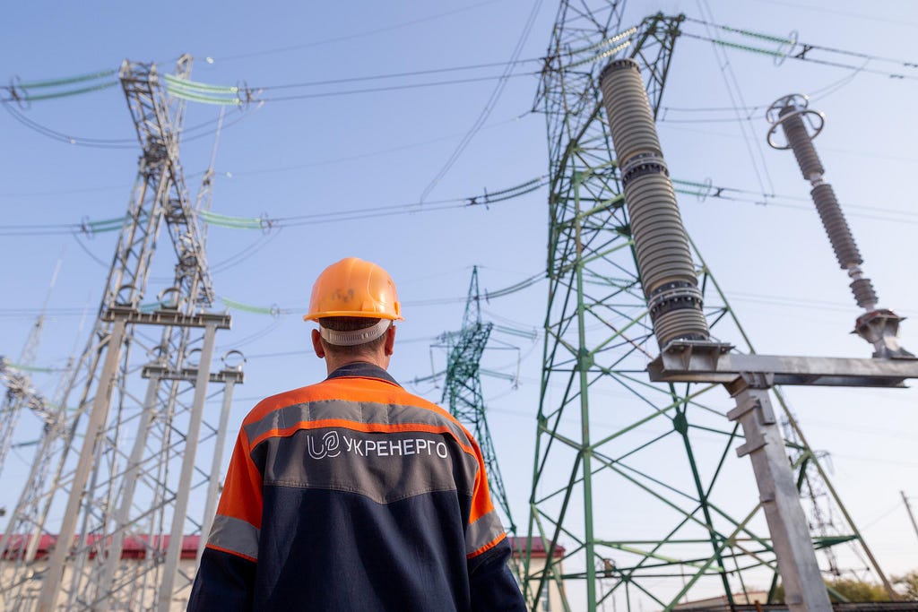 Man wearing hardhat and an orange and blue jacket with writing in Ukrainian that says ‘Ukrenergo’ looks at electricity pylons.
