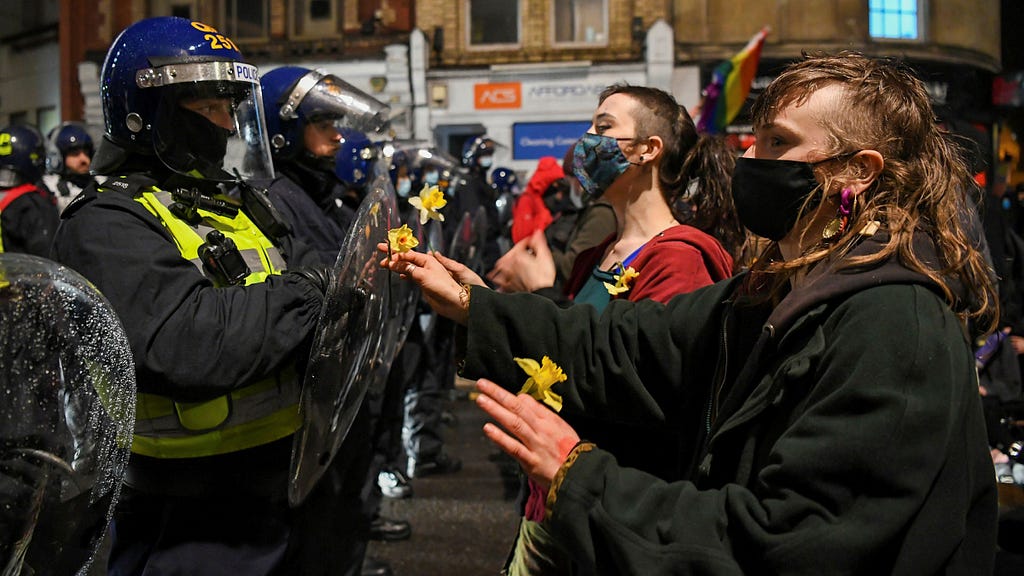Police in riot gear with helmets and plastic shields in confrontation with those on the left of the image: young people wearing face masks. They are outdoors in a street, shops faintly seen in the background.