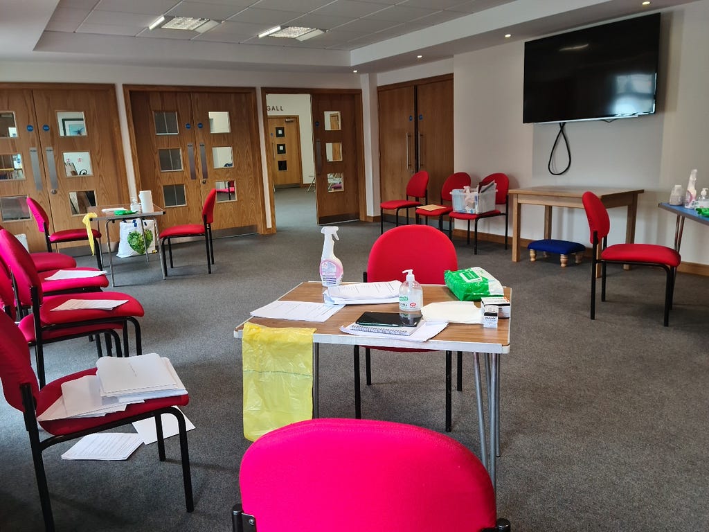 A room with red chairs around the outside of it, some of which have pieces of paper on them. There are three tables that have papers, cleaning spray and bottles of hand sanitizer on them.