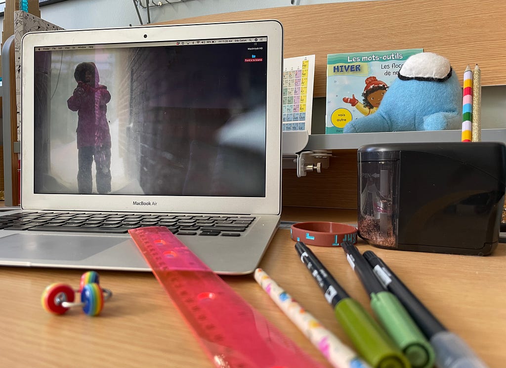 A laptop on a desk with a photo of a child in a storm, yelling on the desktop. There are pens and pencils on the desk, a stim toy and a ruler. In the back is a French book about winter and a stuffed Among Us video game character.