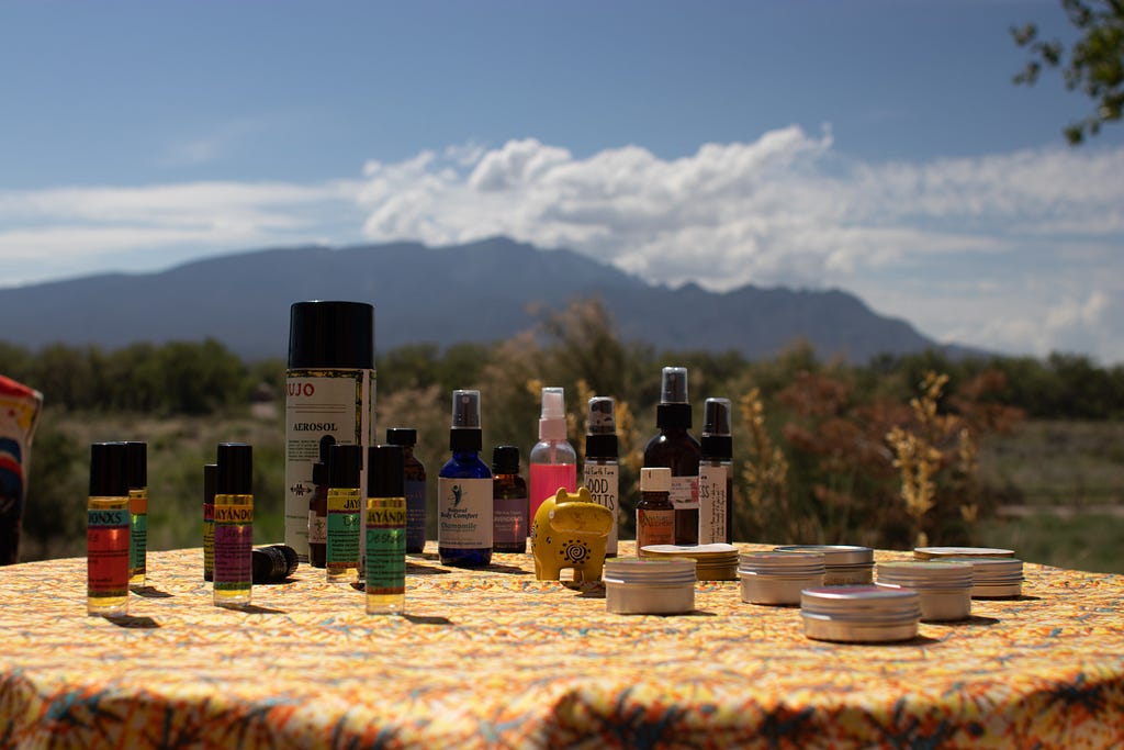 Photograph of multiple tinctures, salves, and sprays on a table, with the Sandia Mountains in the background