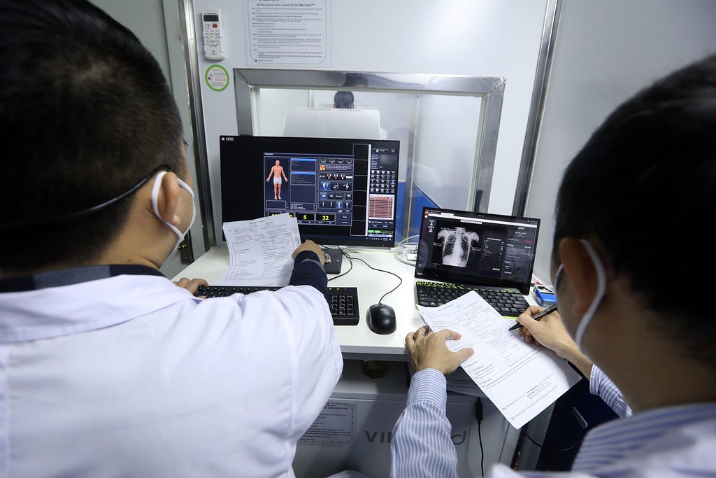 Two health care workers sit at a desk with two computer monitors that show a digital scan of an X-ray and details about the health of the patient.