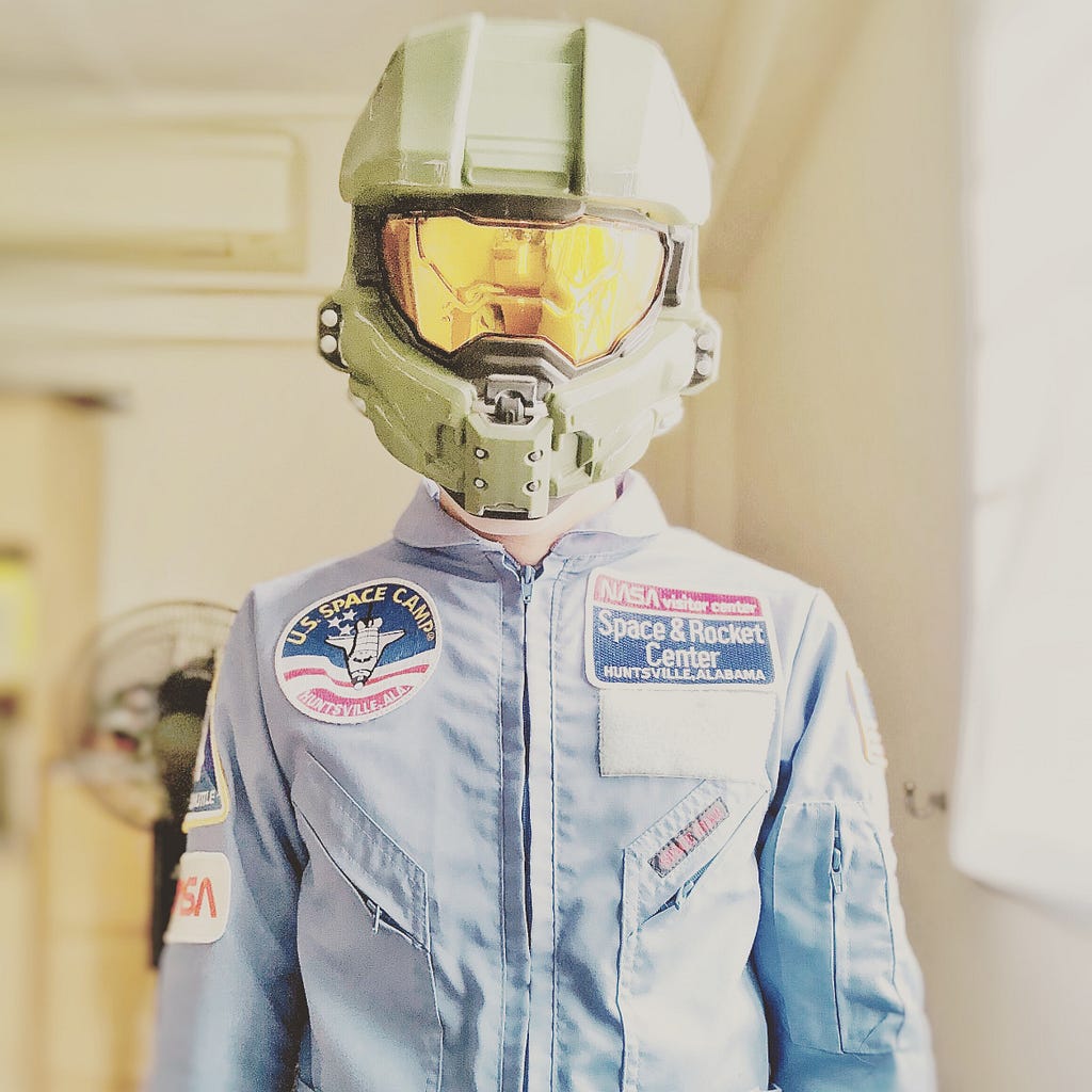 A photo of a person, likely a child, wearing a light-blue US Space Camp jump-suit. On their head, is a helmet from the video-game series Halo.