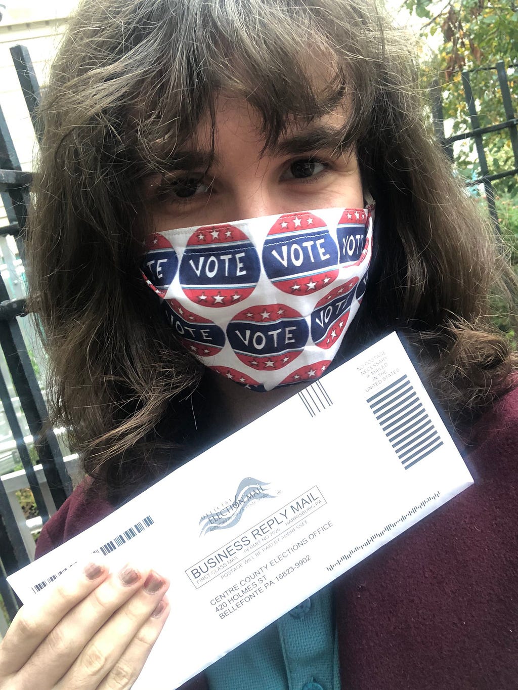 A selfie of the author holding their mail-in ballot envelop and wearing a mask covered in “vote” stickers