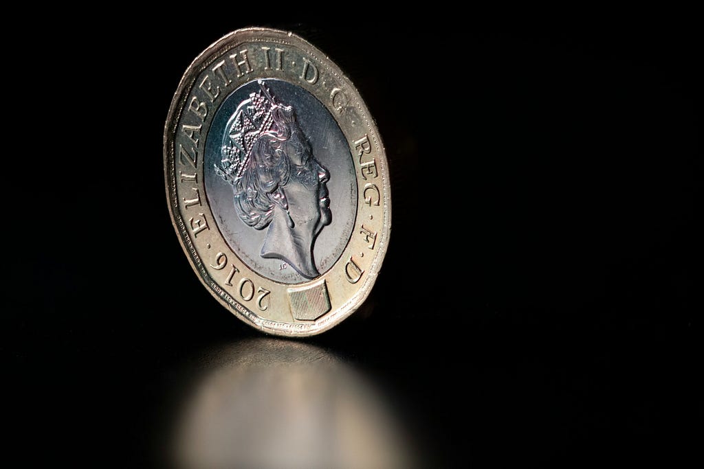 A coin balancing on its edge. The coin is a British pound coin and the  heads side is facing us. The heads side features Queen Elizabeth II’s profile.