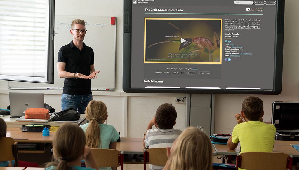 Teacher standing in front of his class and a smartboard that is showing the AI Scene Description tool describing a video about insects.