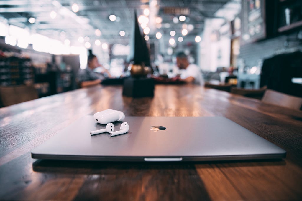 A closed grey MacBook sits on a table in a brightly lit space. A pair of AirPods and their case sit on top.