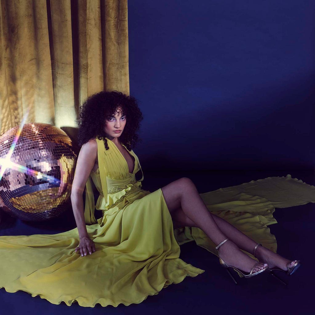 Model in yellow dress sitting against blue backdrop, leaning on glitterball