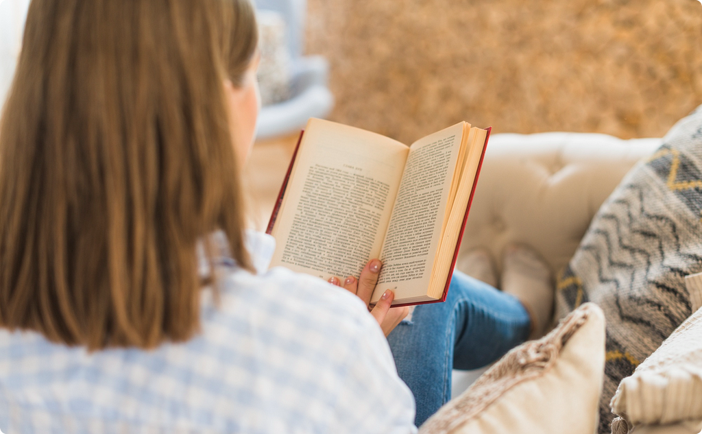 an image of a lady wearing a blue jean and a shirt sitting in a sofa while reading a book