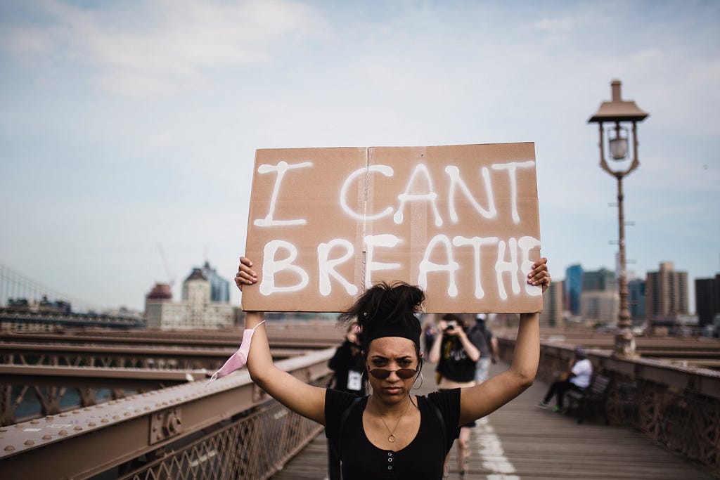 A woman stands on a boardwalk with a cityscape and a large lamppost in the background. She is wearing a black short sleeved shirt and sunglasses that are lowered underneath her eyes. She looks directly into the camera and holds a homemade sign of cardboard over her head. It reads “I CANT BREATHE.”