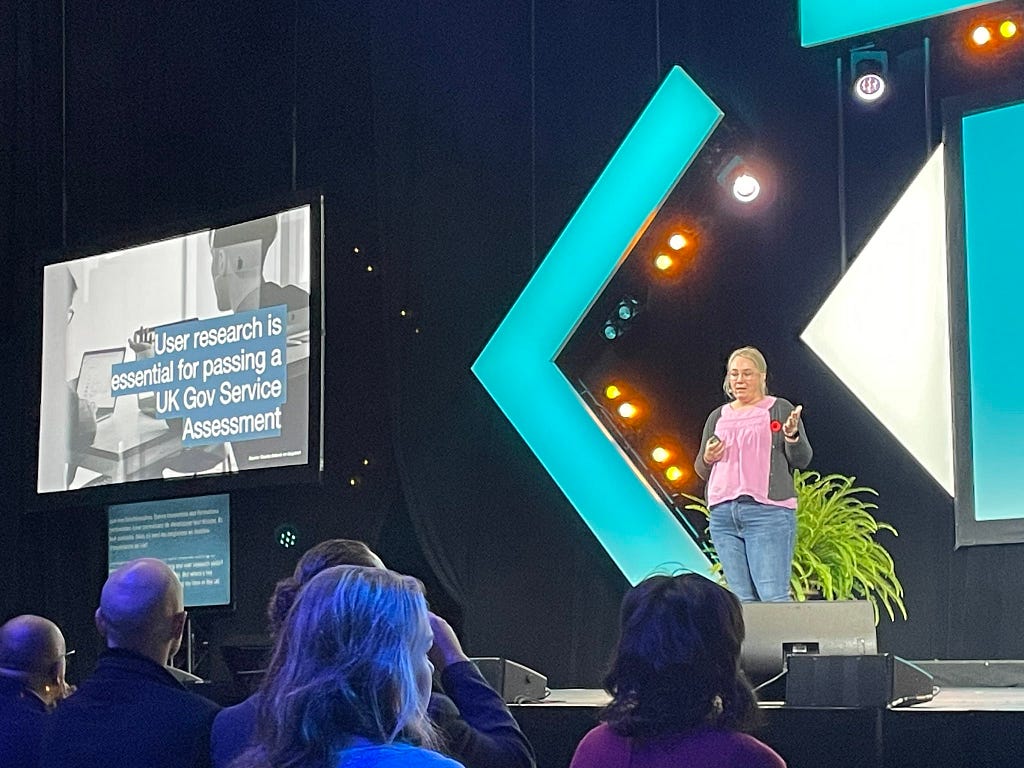 A blonde woman in a purple shirt, black cardigan and jeans, speaking on a stage