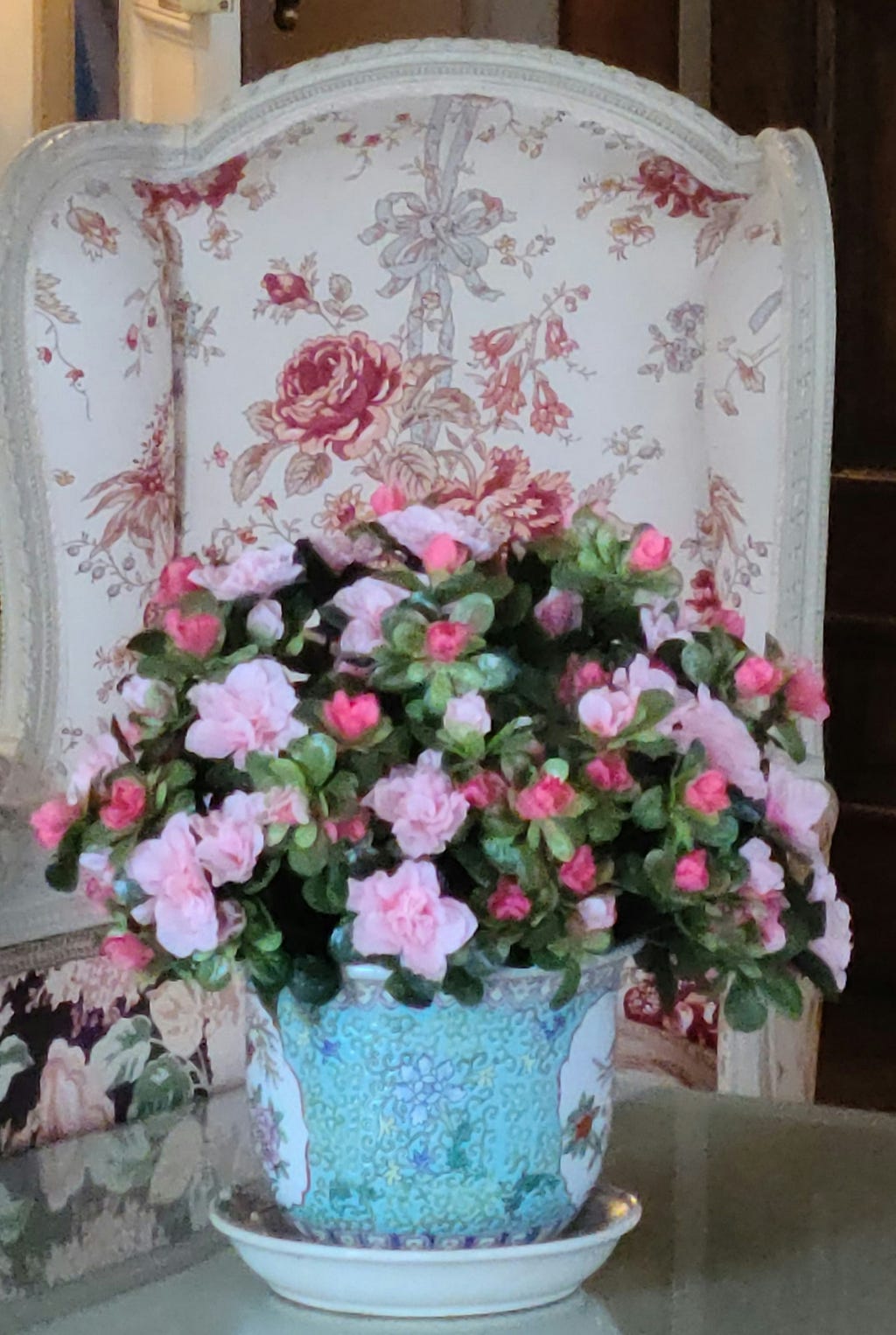 A table arrangement of flowers in various shades of pink in a blue vase on a table with a high backed chair with a floral pattern in rose and pink.
